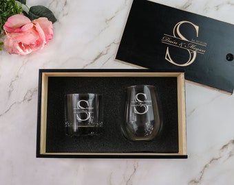 May include: A black wooden box with a black lid. The lid has a gold engraved design with the text "Est. 03.21.20 Dain & Megan". The box contains two clear glasses, one with a whiskey tumbler shape and one with a wine glass shape. Both glasses have a gold engraved design with the letter "S".