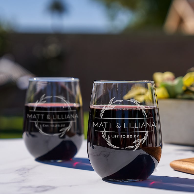 Eye-level view of two engraved stemless wine glasses. The etching has a circular leafy border with the names Matt & Lilliana in the center with Est. 10.25.22 underneath them. The glasses are sitting on a white outdoor table.