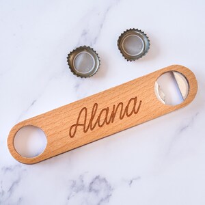 May include: A wooden bottle opener with a metal opener on one end and the name "Alana" engraved on the surface. Two bottle caps are in the background.