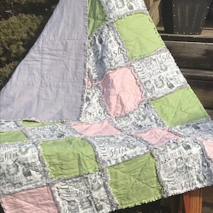 May include: A patchwork quilt with a light gray, pink, and green color scheme. The quilt is made of squares with different patterns and textures. The quilt has a ruffled edge and features the text "Shine bright, little one".