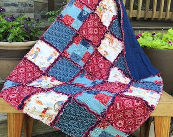May include: A patchwork quilt with a variety of patterned squares in red, blue, and white. The quilt is draped over a wooden bench. The quilt's design includes floral, paisley, and cowboy-themed patterns. The quilt has a dark blue backing.
