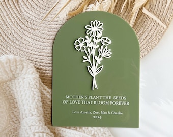May include: A green arch-shaped plaque with a white floral design and the text "Mother's plant the seeds of love that bloom forever. Love Amelia, Zoe, Max & Charlie 2024".