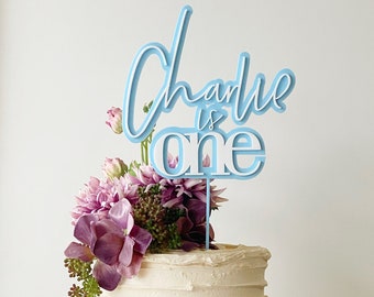May include: A two-tiered white cake decorated with purple flowers and a blue cake topper that says "Charlie is one".
