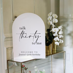 May include: A white arch-shaped sign with black text that reads "talk thirty to me" and "Welcome to Jessica's 30th birthday". The sign is on a gold metal stand with a glass top. White orchids are in a gold vase on the stand. A large bouquet of pink, white, and green flowers is on the floor in front of the stand.