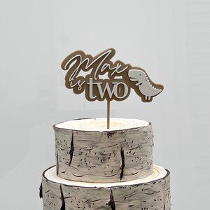 May include: A two-tiered cake decorated with a brown and white birch bark design. A brown and white cake topper with the text "Max is two" and a small white dinosaur is on top of the cake.