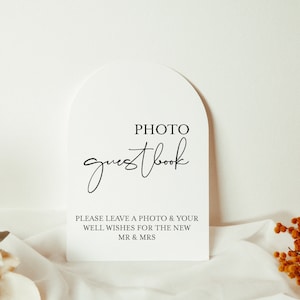 May include: White arch-shaped sign with the text "PHOTO guestbook" in black script font. Below the text is a message that reads "PLEASE LEAVE A PHOTO & YOUR WELL WISHES FOR THE NEW MR & MRS".