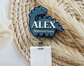 May include: A blue dinosaur-shaped badge holder with the name "ALEX" and the text "Registered Nurse" in white lettering. The badge holder is attached to a white retractable badge reel.