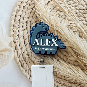 May include: A blue dinosaur-shaped badge holder with the name "ALEX" and the text "Registered Nurse" in white lettering. The badge holder is attached to a white retractable badge reel.