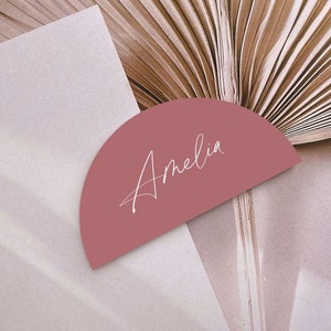 May include: A half-circle shaped place card with the name Amelia written in white cursive on a dusty rose background.