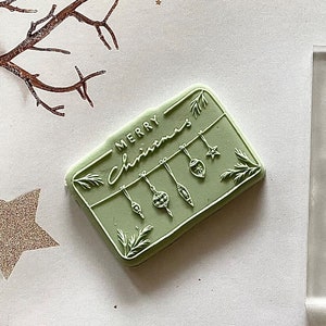 May include: A green rubber stamp with the text "Merry Christmas" and a design of hanging ornaments. The stamp is on a white surface with gold glitter stars.