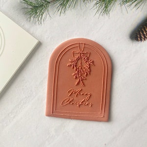 May include: A terracotta-colored rubber stamp with an arched design. The stamp features a Christmas-themed illustration of a sprig of holly with a bow and bells, and the text "Merry Christmas".