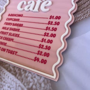 Puede incluir: Un letrero de caf&eacute; rosa y blanco con un borde ondulado. El letrero dice "Bienvenido al caf&eacute; de [Name]" y enumera los precios de varios art&iacute;culos del men&uacute;, que incluyen babycino, cupcake, pan de hadas, batido de leche, rodajas de fruta, helado, galletas y tostadas.