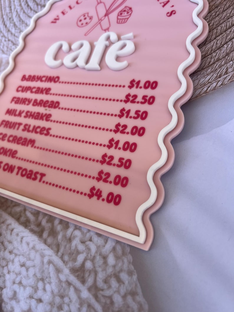 Puede incluir: Un letrero de caf&eacute; rosa y blanco con un borde ondulado. El letrero dice "Bienvenido al caf&eacute; de [Name]" y enumera los precios de varios art&iacute;culos del men&uacute;, que incluyen babycino, cupcake, pan de hadas, batido de leche, rodajas de fruta, helado, galletas y tostadas.