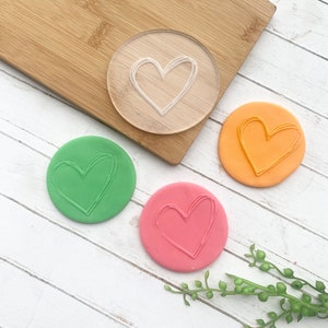 Puede incluir: Tres piezas redondas de plastilina con una forma de coraz&oacute;n impresa en cada una. La plastilina es verde, naranja y rosa. Tambi&eacute;n se muestra un sello de coraz&oacute;n de pl&aacute;stico transparente.