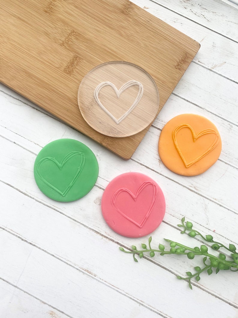 Puede incluir: Tres piezas redondas de plastilina con una forma de coraz&oacute;n impresa en cada una. La plastilina es verde, naranja y rosa. Tambi&eacute;n se muestra un sello de coraz&oacute;n de pl&aacute;stico transparente.