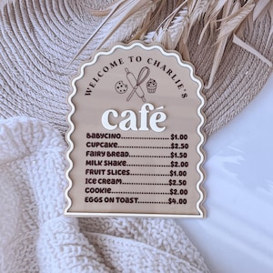 Puede incluir: Un letrero de madera marr&oacute;n con un borde blanco festoneado y el texto "Bienvenido al caf&eacute; de Charlie" en blanco. El letrero enumera los precios de varios art&iacute;culos de caf&eacute;, que incluyen babycino, cupcake, pan de hadas, batido de leche, rodajas de fruta, helado, galleta y huevos con tostadas.