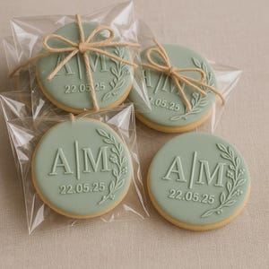 May include: Four round cookies with a pale green icing, individually wrapped in clear plastic. The cookies display the initials "AM" and the date "22.05.25" with a floral design. Each cookie is tied with twine.