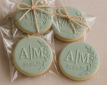 May include: Four round cookies with a light green frosting, each individually wrapped in clear plastic. The cookies feature the initials "AM" and the date "22.05.25" with a floral design. Each cookie is tied with twine.