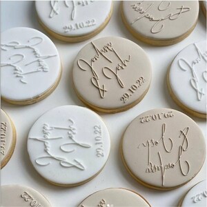 May include: Round cookies with white and tan icing. Each cookie is decorated with the names "Laura & Josh" and the date "29.10.22" in an elegant script. The cookies have a light brown edge.