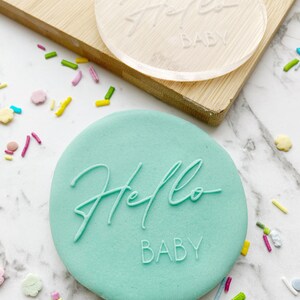 May include: A light green round cookie with the words "Hello Baby" embossed on it. The cookie is surrounded by colorful sprinkles.