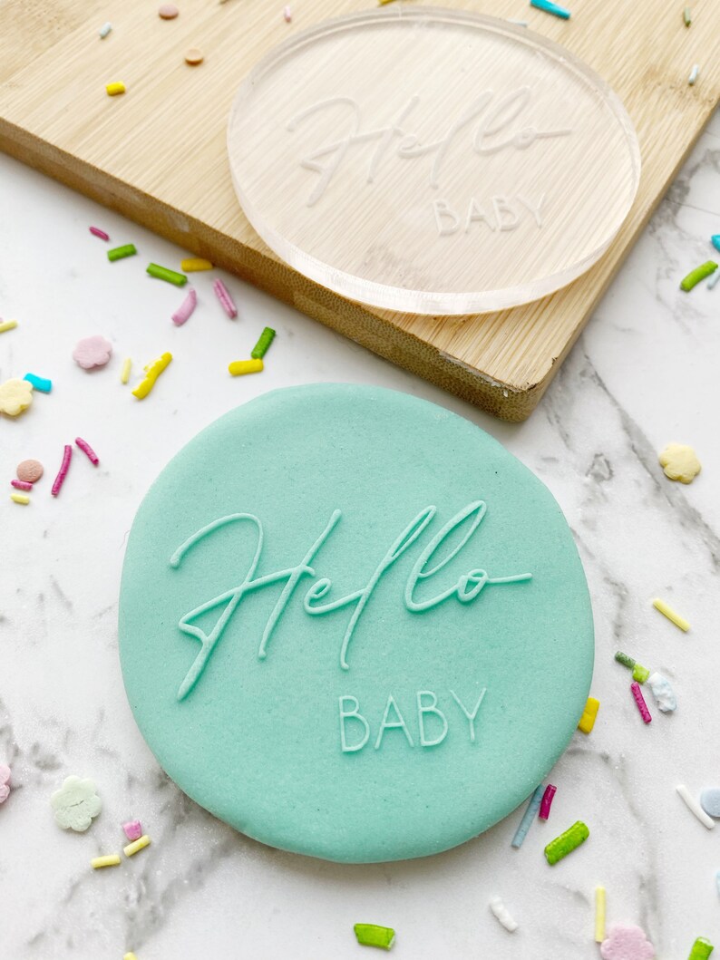 May include: A light green round cookie with the words "Hello Baby" embossed on it. The cookie is surrounded by colorful sprinkles.