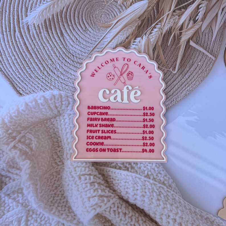Puede incluir: Un letrero de madera rosa y blanco con un borde festoneado que dice "Bienvenido al caf&eacute; de Cara" y enumera los precios de varios art&iacute;culos del men&uacute;, que incluyen babycino, cupcake, pan de hadas, batido de leche, rodajas de fruta, helado, galleta y huevos con tostadas.