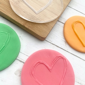 Puede incluir: Tres piezas redondas de plastilina con una forma de coraz&oacute;n impresa en cada una. La plastilina es verde, naranja y rosa. Un sello de pl&aacute;stico transparente en forma de coraz&oacute;n est&aacute; sobre una tabla de cortar de madera.