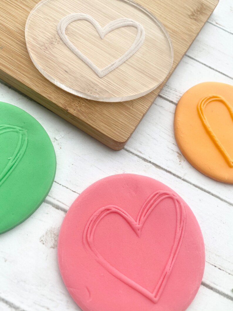 Puede incluir: Tres piezas redondas de plastilina con una forma de coraz&oacute;n impresa en cada una. La plastilina es verde, naranja y rosa. Un sello de pl&aacute;stico transparente en forma de coraz&oacute;n est&aacute; sobre una tabla de cortar de madera.