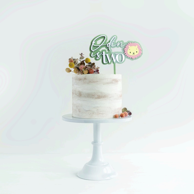 May include: A white frosted cake with dried flowers on top, displayed on a white cake stand. A cake topper reads "Odin is two" with a lion illustration. The cake has a rustic, semi-naked design.
