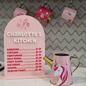 Pode incluir: Uma placa rosa com o texto "Charlotte's Kitchen" e um menu de itens com pre&ccedil;os. O menu inclui babycino, cupcake, p&atilde;o de fada, milk shake, fatias de fruta, sorvete, biscoitos e ovos com torradas. Um regador rosa com um design de unic&oacute;rnio est&aacute; em primeiro plano.