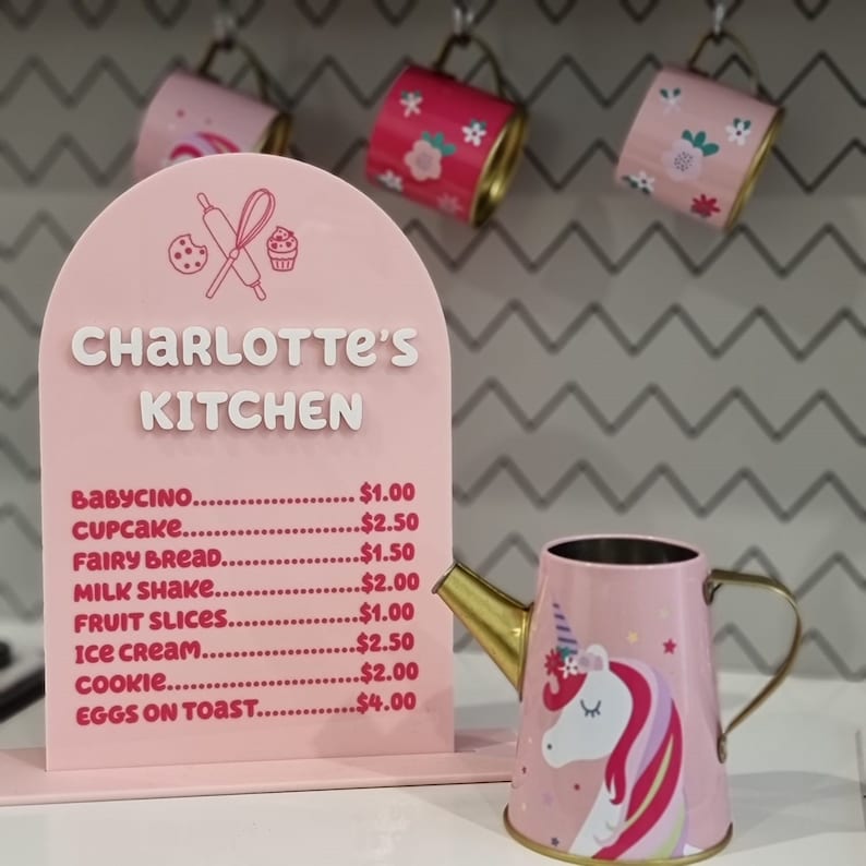Pode incluir: Uma placa rosa com o texto "Charlotte's Kitchen" e um menu de itens com pre&ccedil;os. O menu inclui babycino, cupcake, p&atilde;o de fada, milk shake, fatias de fruta, sorvete, biscoitos e ovos com torradas. Um regador rosa com um design de unic&oacute;rnio est&aacute; em primeiro plano.