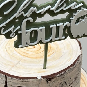 May include: Green and white cake topper with the text "Charlie is four" and a dinosaur silhouette. The topper is on a wooden cake stand.