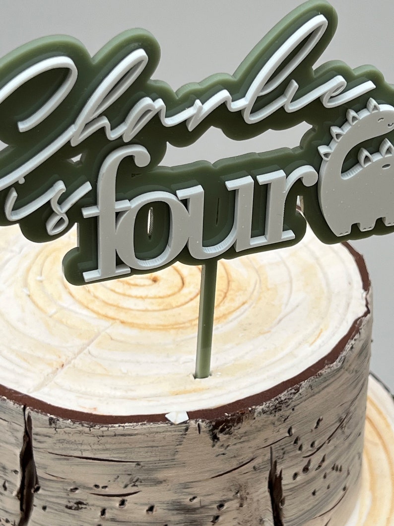 May include: Green and white cake topper with the text "Charlie is four" and a dinosaur silhouette. The topper is on a wooden cake stand.
