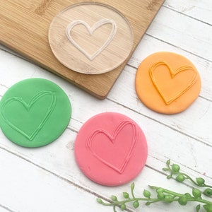 Puede incluir: Un sello de pl&aacute;stico transparente en forma de coraz&oacute;n con una impresi&oacute;n en forma de coraz&oacute;n verde, naranja y rosa sobre una superficie de madera blanca.