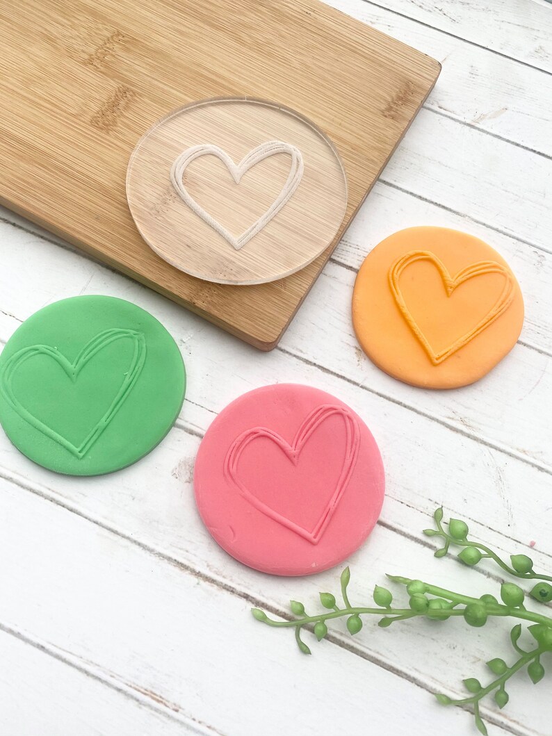 Puede incluir: Un sello de pl&aacute;stico transparente en forma de coraz&oacute;n con una impresi&oacute;n en forma de coraz&oacute;n verde, naranja y rosa sobre una superficie de madera blanca.
