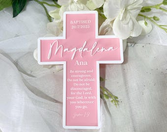 Puede incluir: Una cruz rosa para bautizo con borde blanco. La cruz presenta el nombre "Magdalena" en cursiva, el nombre "Ana" y la fecha "20/7/2025". También se muestra el texto "Sé fuerte y valiente..."