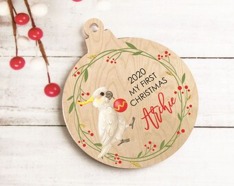 Op de afbeelding: Een houten kerstversiering met een witte kaketoe die een rode en gele bal vasthoudt. De versiering is versierd met een groene krans met rode bessen en gouden accenten. De tekst "2020 My First Christmas Archie" is op de versiering gedrukt.