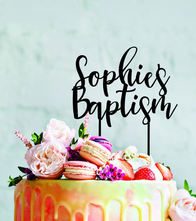 May include: Black acrylic cake topper with the text "Sophie's Baptism" in a cursive font. The topper is on a white cake decorated with pink and yellow frosting, macarons, strawberries, and flowers.