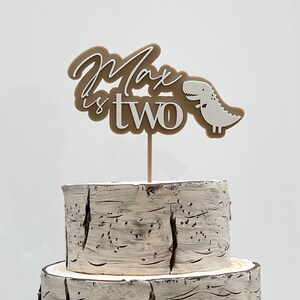 May include: A brown and white cake topper with the text "Max is two" and a small white dinosaur. The topper is on a two-tiered cake with a birch bark design.