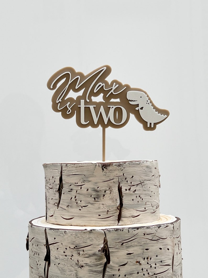 May include: A brown and white cake topper with the text "Max is two" and a small white dinosaur. The topper is on a two-tiered cake with a birch bark design.