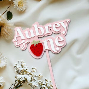 May include: A pink and white cake topper with the text "Aubrey is one" and a red strawberry illustration. The topper is on a pink stick, ideal for a first birthday celebration.