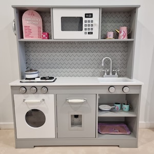 Pode incluir: Uma cozinha de brincar de madeira cinza com um forno, um forno de microondas e um fog&atilde;o brancos. A cozinha tem uma bancada cinza, uma pia e duas prateleiras. A cozinha tem uma placa que diz "Charlotte's Kitchen" e um menu com pre&ccedil;os para diferentes itens de comida.