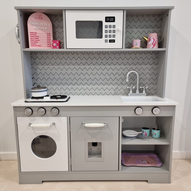 Pode incluir: Uma cozinha de brincar de madeira cinza com um forno, um forno de microondas e um fog&atilde;o brancos. A cozinha tem uma bancada cinza, uma pia e duas prateleiras. A cozinha tem uma placa que diz "Charlotte's Kitchen" e um menu com pre&ccedil;os para diferentes itens de comida.