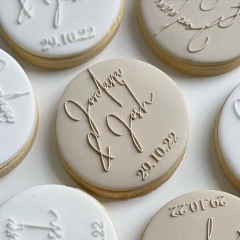 May include: Close-up of several round cookies with white and tan icing. The cookies are decorated with names and dates in elegant script. The cookies are arranged on a white surface, with a few in focus.