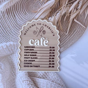 Puede incluir: Un letrero de madera con borde festoneado y fondo marr&oacute;n. El letrero dice "Bienvenido al caf&eacute; de Charlie" y enumera los precios de varios art&iacute;culos del men&uacute;, incluyendo babycino, cupcake, pan de hadas, batido de leche, rodajas de fruta, helado, galleta y huevos con tostadas.