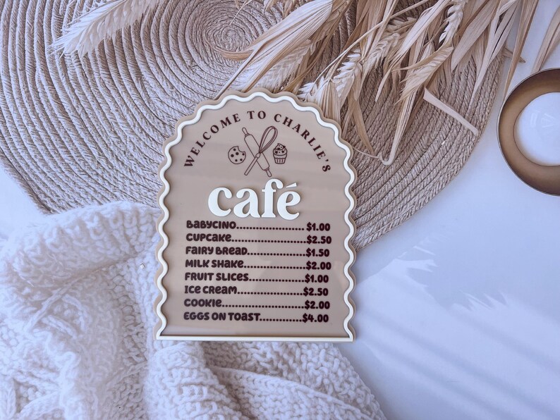 Puede incluir: Un letrero de madera con borde festoneado y fondo marr&oacute;n. El letrero dice "Bienvenido al caf&eacute; de Charlie" y enumera los precios de varios art&iacute;culos del men&uacute;, incluyendo babycino, cupcake, pan de hadas, batido de leche, rodajas de fruta, helado, galleta y huevos con tostadas.