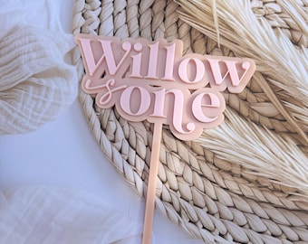 May include: Pink acrylic cake topper with the text "Willow is one" in a cursive font. The topper is on a woven straw background.