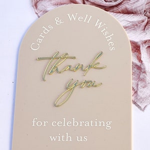 K&ouml;nnte beinhalten: Ein beigefarbenes Schild in Bogenform mit dem Text "Cards & Well Wishes" oben und "Thank You for celebrating with us" unten. Das Schild hat ein goldenes "Thank You" in kursivschrift.