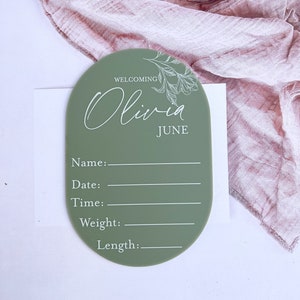 May include: A green oval-shaped acrylic sign with a floral design and the text "Welcoming Olivia June". The sign has blank lines for the baby's name, date, time, weight, and length.