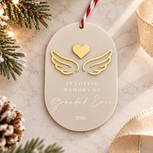 May include: An oval-shaped beige acrylic ornament featuring a gold-colored heart and angel wings. The text reads IN LOVING MEMORY OF Grandad Eric 2026. A red and white striped string is attached to the top for hanging the decorative piece.